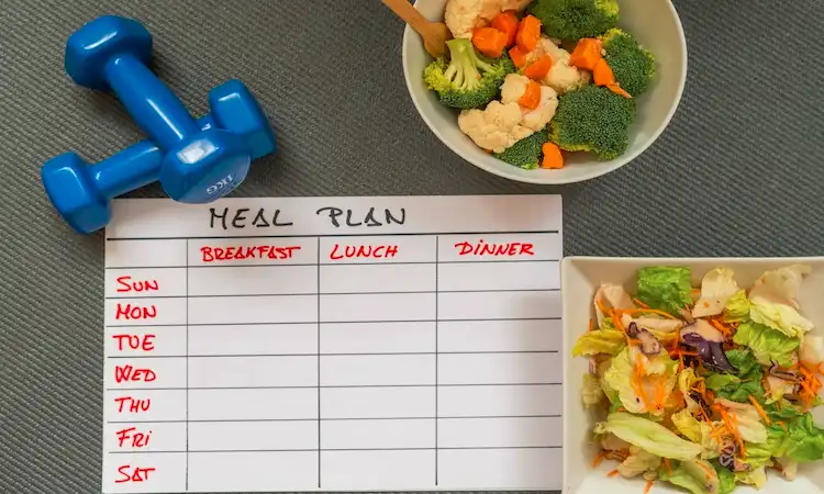 NEAT Calories, the Fat Loss Lever You Miss A meal plan chart with days and meal sections is placed next to a blue dumbbell, a bowl of mixed vegetables, and a salad. The tone conveys health and organization.