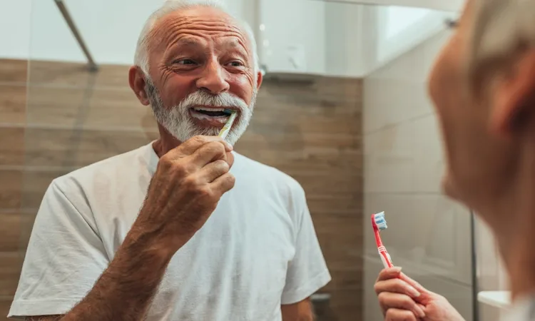 Oral Health for Longevity Blueprint Elderly man smiling while brushing teeth in a bathroom, wearing a white T-shirt. Cheerful atmosphere, woman in foreground holds a toothbrush, about oral health for longevity.