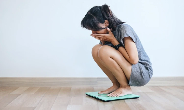 Stress and Weight Gain Biology Facts A woman crouches on a scale in a minimalist room, covering her face with hands, conveying shock or distress. She wears a gray dress, with a neutral backdrop.
