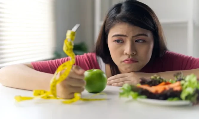 A frustrated young woman looking at a green apple and salad while holding a yellow measuring tape and think why weight loss stops after losing 20 pounds