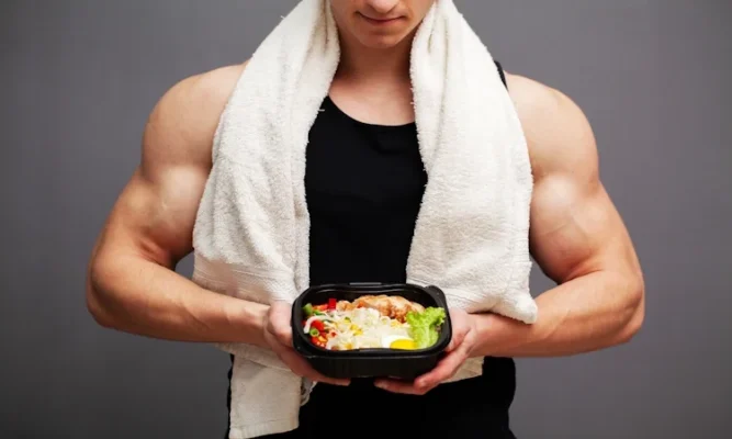 A muscular man with a white towel draped over his shoulders holding a black meal prep container filled with chicken, a hard-boiled egg, and vegetables for protect muscle while cutting calories