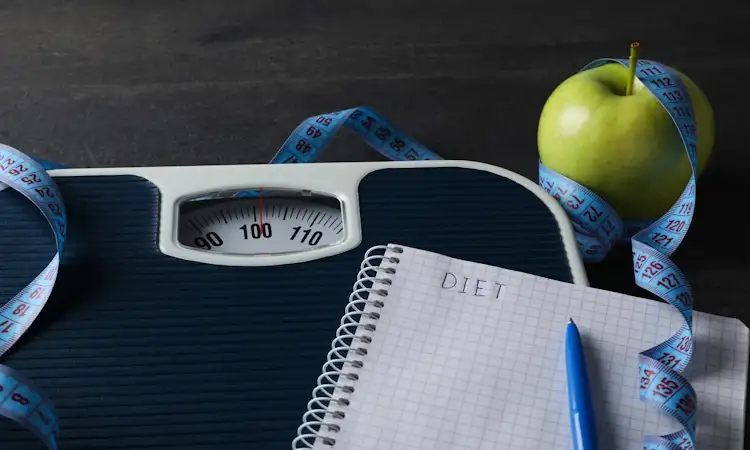 A bathroom scale showing approximately 100 kilograms, next to a green apple wrapped in a blue measuring tape, and a notebook with the word 'DIET' written on it for set point theory and weight loss resistance.