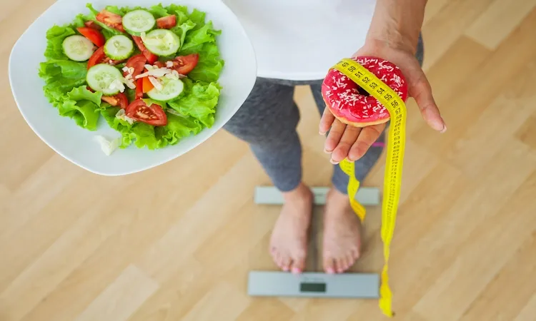 An overhead view of a woman standing on a glass scale, holding a fresh green salad in one hand and a pink frosted donut wrapped in a yellow measuring tape in the other, about metabolic adaptation and weight loss.