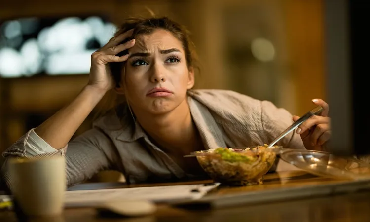 A frustrated woman looking exhausted and asking 'why do I feel tired and inactive while dieting?' as she stares at a computer screen. She is resting her head on her hand with a stressed expression while eating a salad during a late-night study or work session at her desk.