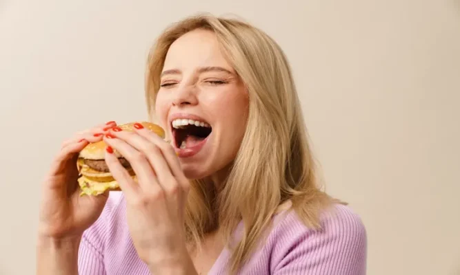 A happy blonde woman with red nails laughing and opening her mouth wide to take a big bite of a juicy double cheeseburger and think Why hunger hormones increase after calorie deficit.