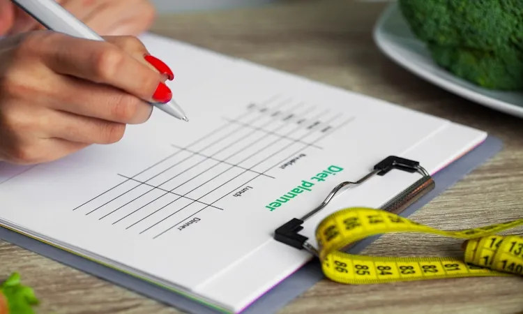 An overhead, close-up shot shows a troubleshooting a fat loss plateau checklist with red-manicured nails writing on a Diet Planner chart resting on a clipboard. A yellow measuring tape and a plate of fresh broccoli are visible on the wooden table, symbolizing health monitoring and clean eating.