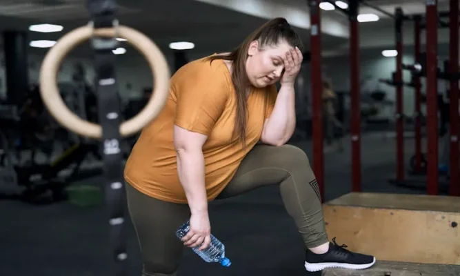 Resting metabolic rate after weight loss. An overweight woman stands in a gym, leaning forward with one foot resting on a wooden plyometric box. She looks exhausted, holding a bottle of water in one hand and resting her other hand against her forehead as she catches her breath. In the blurred background, gymnastics rings and gym equipment are visible, emphasizing a high-intensity workout environment.