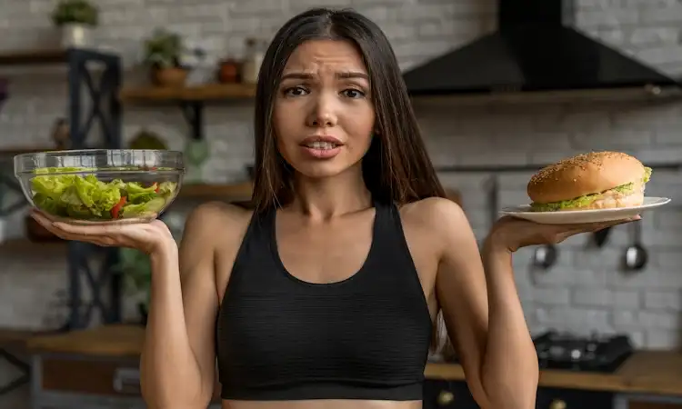 A young woman in athletic wear holding a fresh green salad in one hand and a hamburger in the other, looking conflicted about a food choice and think what are the signs of metabolic adaptation from dieting